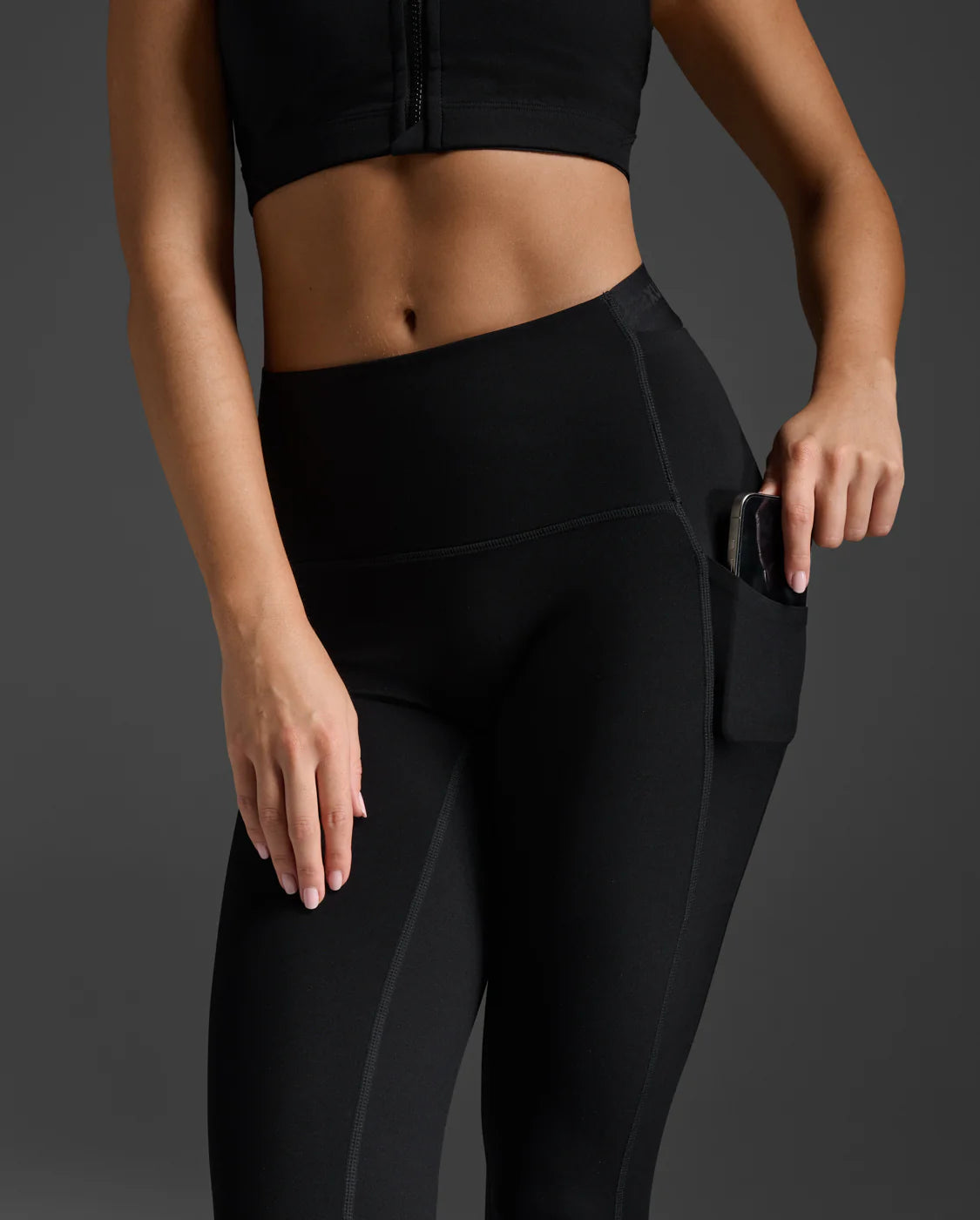 Side view of woman wearing black 2XU Form Stash compression leggings highlighting the deep side hip pocket for smartphone storage.