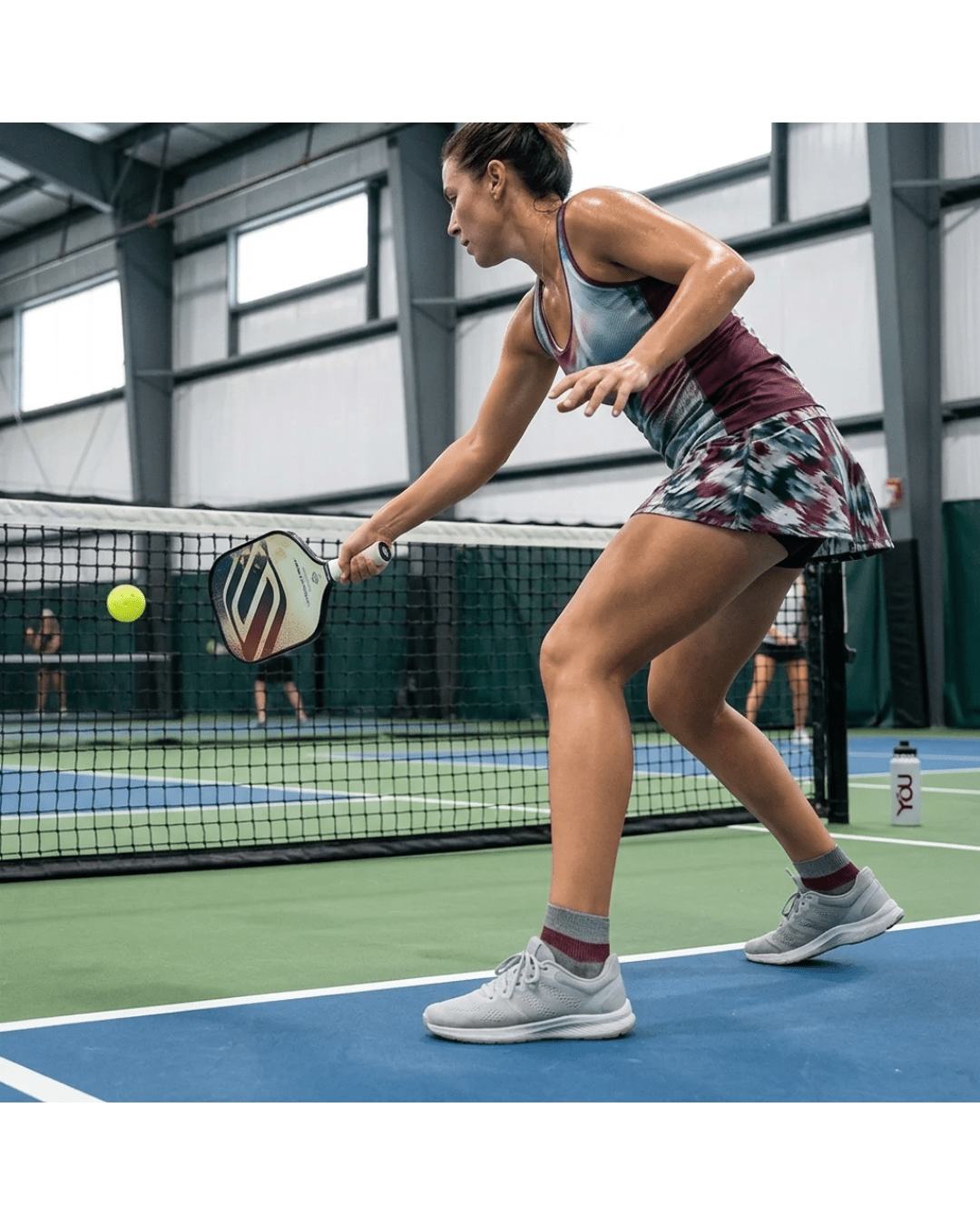 A female athlete playing pickleball on an indoor court wearing YoU Compression® Grey &amp; Burgundy 20-30 mmHg Ankle Socks with light gray athletic shoes.
